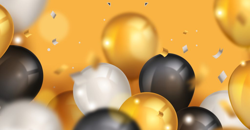 A képen nagyméretű, fényes felületű arany-, fekete- és fehér lufik láthatók zsúfoltan egymás mellett. A háttér egységes, meleg narancssárga színű. A levegőben apró arany és ezüst konfettidarabok úsznak, mintha egy ünneplés vagy rendezvény pillanatát örökítették volna meg. A lufik fényvisszaverődései és a lágy elmosódások dinamikus, mozgalmas hatást keltenek.